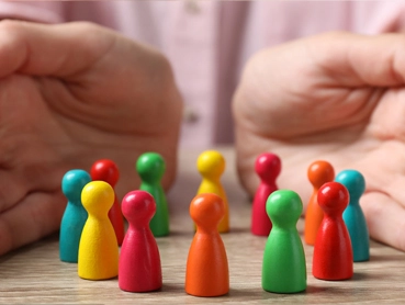 A pair of hands encircles a group of colorful, upright game pieces on a wooden surface. The pieces are painted in various bright colors, including red, green, yellow, and blue, representing people in a protected environment.
