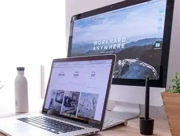 A home office setup features an open laptop and a desktop monitor displaying different screens. The desktop monitor shows the text "WORK HARD ANYWHERE" over a scenic background. A water bottle and a pen in a holder are placed beside the laptop.
