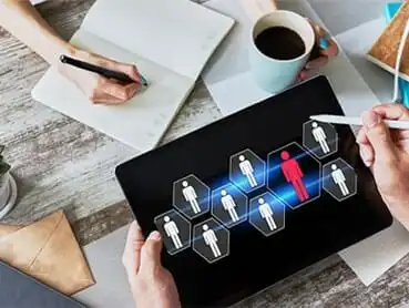 A top-down view shows a tablet displaying a network diagram with multiple human icons, one highlighted in red. Nearby, a person writes in a notebook while another holds a cup of coffee. Various office supplies are scattered on the wooden surface.