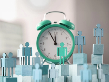 Several light blue stick-figure cutouts stand on variously sized wooden blocks in the foreground. A green retro-style alarm clock displays the time at around 8:00 in the background, indicating a conceptual scene likely representing time to hire, time management, or teamwork.