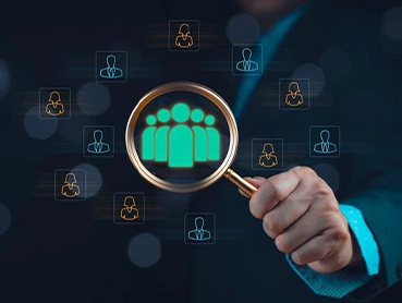 A person holds a magnifying glass highlighting a group of three green, abstract human figures, symbolizing enterprise talent acquisition. Surrounding the magnifying glass are icons of individual avatars displayed in blue and yellow against a dark background.
