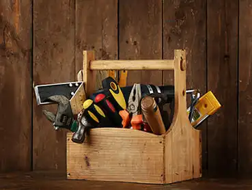 A wooden toolbox with a handle contains various tools, including a handsaw, screwdrivers, pliers, a wrench, a measuring tape, and other hand tools. The background is made of rustic wooden planks.