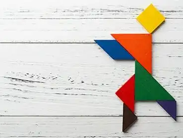 A colorful tangram puzzle piece arrangement forming the shape of a person running against a white wooden background. The pieces include a yellow square for the head, orange and red triangles for the body, and blue, green, and brown shapes for the arms and legs.