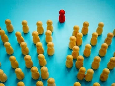 A red game piece stands apart from a crowd of identical yellow game pieces on a blue background, symbolizing individuality or leadership.