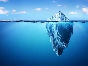 A large iceberg floating in the middle of a deep blue ocean. The tip of the iceberg is visible above the water, while a massive, jagged section extends below the surface. The sky above is clear with a few scattered clouds.