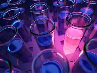 Rows of transparent test tubes, each containing a blue liquid, are arranged closely together. One test tube stands out with a glowing pink liquid, set against a background illuminated in shades of purple and blue.