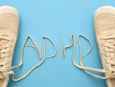 Two tan sneakers are positioned on a light blue background. The shoelaces are untied and arranged to spell "ADHD" between the shoes.