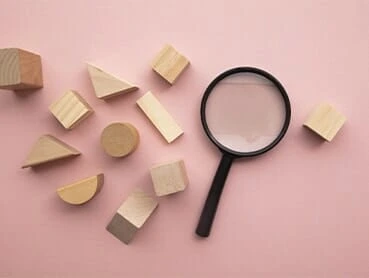 A magnifying glass lies on a pink background surrounded by various wooden geometric shapes, including cubes, cylinders, triangles, and semi-circles.