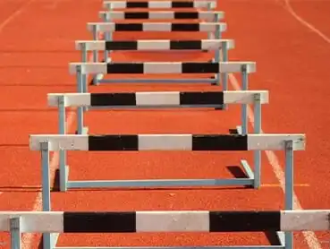 A row of black and white hurdles arranged on an orange running track, stretching into the distance. The track has white lines marking lanes, and the hurdles are evenly spaced, ready for a race.