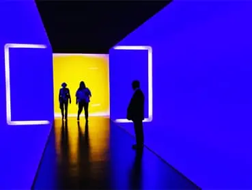 A narrow hallway with vivid blue walls and a bright yellow lit background. Three silhouettes of people are visible: two walking towards the yellow light and one standing to the side, observing. The scene has a modern, futuristic ambiance.