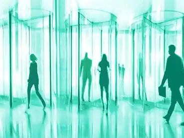 Silhouettes of people walking through a series of transparent, cylindrical revolving doors. The image has a greenish tint, with reflections visible on the shiny floor. Some individuals carry briefcases, implying a business setting.