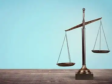 A bronze balance scale stands on a wooden surface against a plain blue background. One side of the scale is higher than the other, indicating an imbalance.