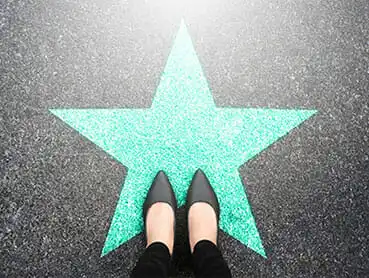 A person wearing black pointed-toe shoes stands on a green star shape painted on a grey pavement, with the star glowing brightly. The image is taken from a top-down perspective.