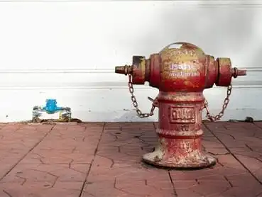 A red fire hydrant is situated on a brick sidewalk against a white wall. The hydrant shows signs of rust and wear. A blue valve or smaller hydrant can be seen in the lower left corner, attached to the wall.