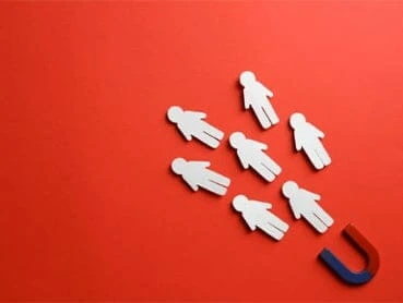 A blue and red horseshoe magnet is attracting six white human figure cutouts on a bright red background, symbolizing the concept of attracting people or customers.