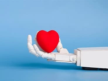 A robotic hand is holding a red heart-shaped object against a plain blue background. The image symbolizes the integration of technology and emotion, suggesting concepts such as artificial intelligence with human-like qualities, or care through technology.