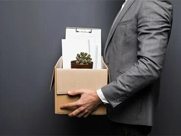 A person in a gray suit holds a cardboard box containing office supplies, including a potted plant, clipboard, and papers. The person is partially visible against a plain dark background.