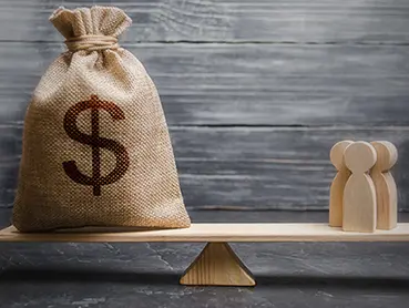 A burlap sack with a large dollar symbol is on one side of a seesaw, while three wooden figurines stand on the other side. The seesaw, balanced on a triangular fulcrum, illustrates the concept of financial balance. The background is wooden planks.