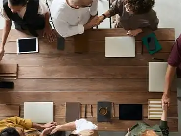 A top-down view of a wooden table with six people gathered around it. Laptops, tablets, and notebooks are placed on the table. Several individuals are shaking hands, indicating a meeting or agreement.