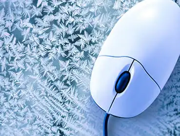 A white computer mouse with a blue cord lies on a frosty, ice-covered surface. The ice forms intricate, crystalline patterns around the mouse, creating a striking contrast between the cold texture of the ice and the smooth plastic finish of the mouse.