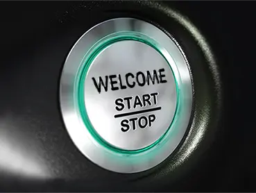 Close-up view of a circular button with a metallic finish. The button is illuminated with a green ring and has the text "WELCOME" at the top, followed by "START" and "STOP" separated by a horizontal line. The button is set against a dark background.