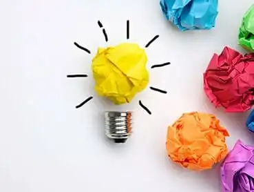 A creative concept image features a lightbulb with a yellow crumpled paper as the bulb, surrounded by crumpled papers in blue, pink, green, red, and orange. Black lines emanate from the yellow paper, symbolizing light.