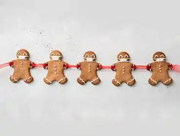 Five gingerbread men cookies decorated with icing are linked together by pieces of red ribbon. The cookies have smiling faces, buttons on their chests, and are placed against a light, plain background.
