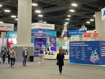 A bustling expo hall filled with people walking and interacting with exhibitor booths from companies like Doccheq.com. Various colorful signs and displays are visible, promoting different tech and business solutions under a large ceiling of industrial lights.