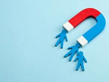 Illustration of a red and blue horseshoe magnet attracting three blue stick figure cutouts, symbolizing the concept of attraction or influence over people. The background is light blue.