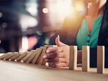 A person wearing a teal shirt and black blazer stops the fall of a line of wooden dominoes with their hand. The background is blurred, with lights creating a bokeh effect.