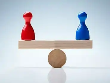 Two game pieces, one red and one blue, are placed on opposite ends of a wooden plank, which is balanced on a cylindrical pivot in the middle, resembling a seesaw. The background is a light gradient blue.