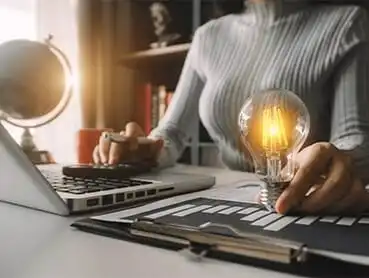 A person sits at a desk working on a laptop, with a small light bulb illuminating next to them. The hand is holding a calculator and there’s a clipboard with a graph on the desk. A globe and bookshelf are in the background, lit by sunlight.